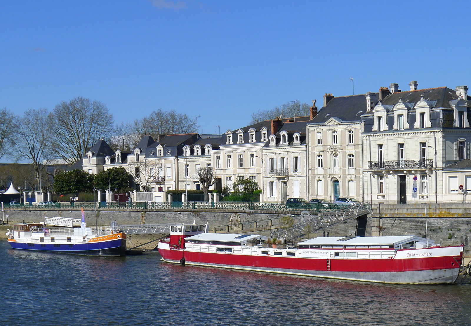 Le quai des Carmes à Angers, dans le quartier de la Doutre : deux péniches amarrées le long d'un quai de pierre au bord de la Maine, maisons angevines claires en arrière-plan