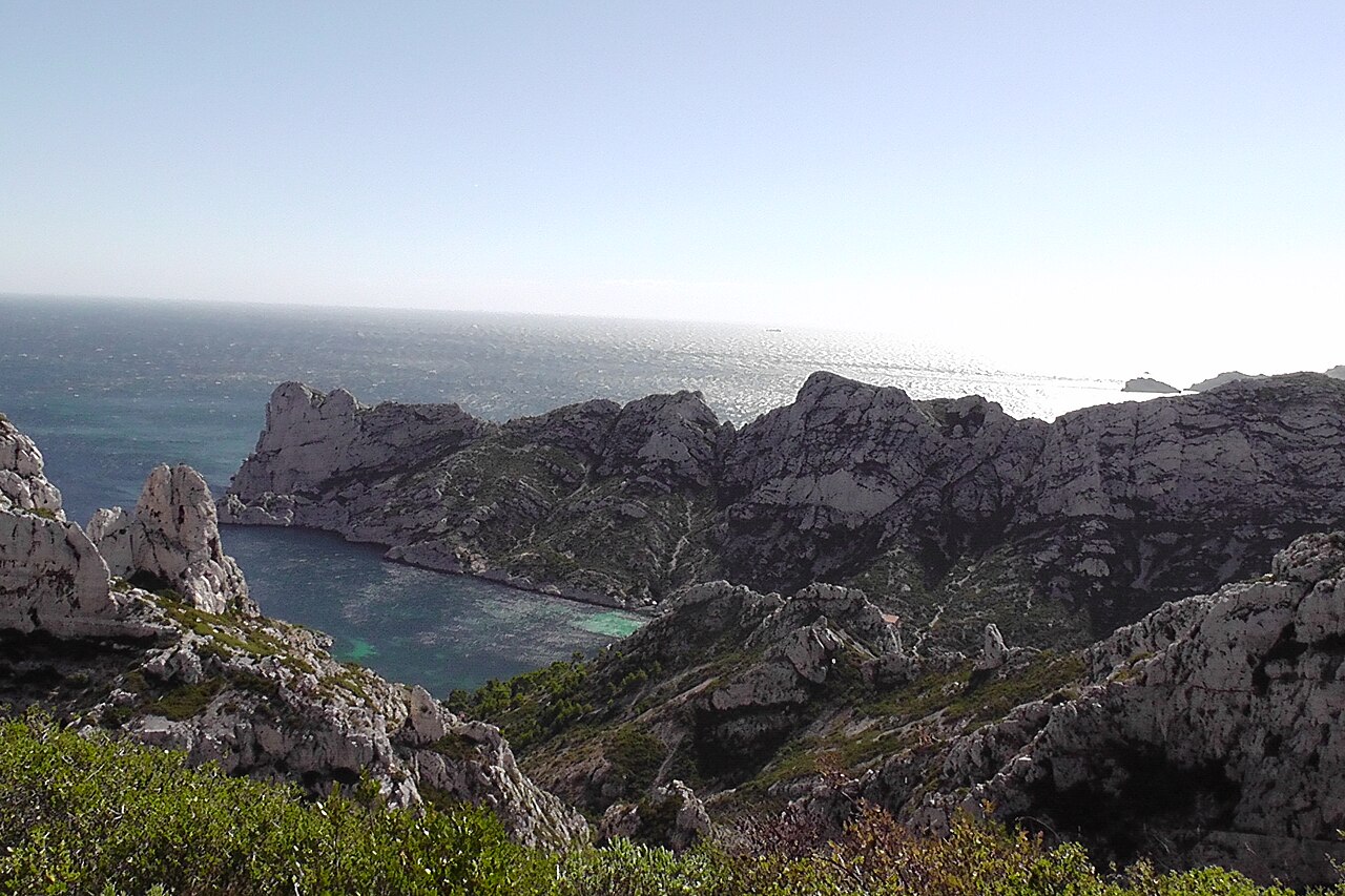 La calanque de Sormiou vue d'en haut : eau turquoise dans une anse fermée par des falaises calcaires blanches, garrigue au premier plan, mer ouverte scintillante à l'horizon