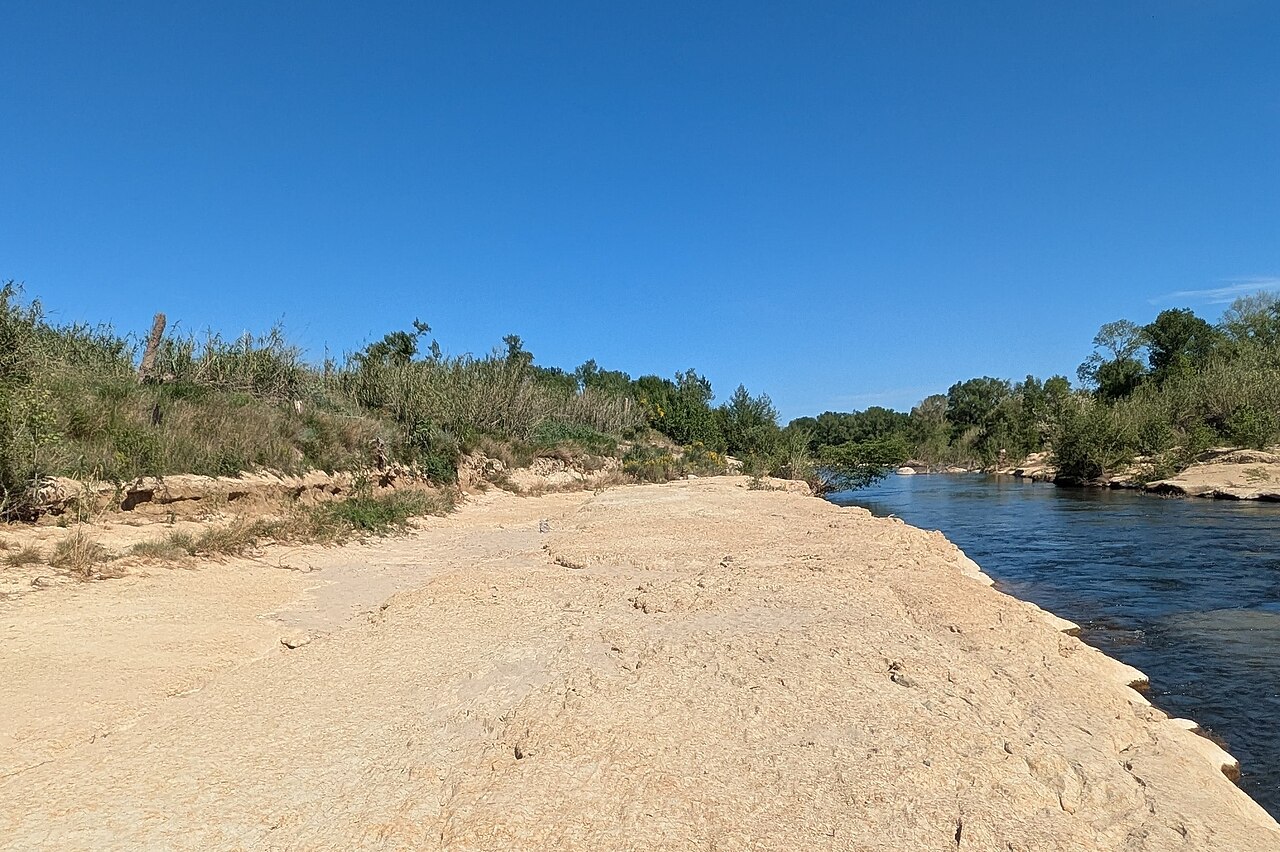 La Têt à l'extrémité ouest de Perpignan : dalle rocheuse claire découverte par l'étiage, filet d'eau bleue à droite, végétation de ripisylve sur les berges, ciel méditerranéen dégagé