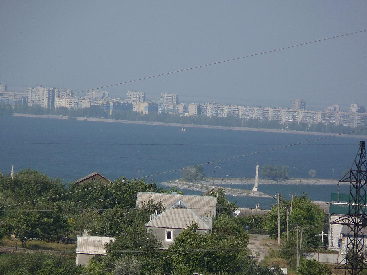 Дніпро біля Кам'янського: широка гладь водосховища, на дальньому березі смуга міських багатоповерхівок, на ближньому приватні будинки й сади, кам'яна дамба з невеликим маяком, лінії електропередач