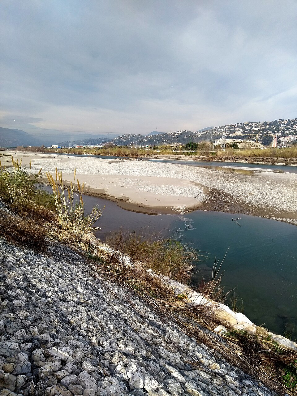 Le lit du Var en basse vallée : larges bancs de galets clairs entre plusieurs bras d'eau peu profonds, enrochement de berge au premier plan, collines urbanisées à l'arrière-plan