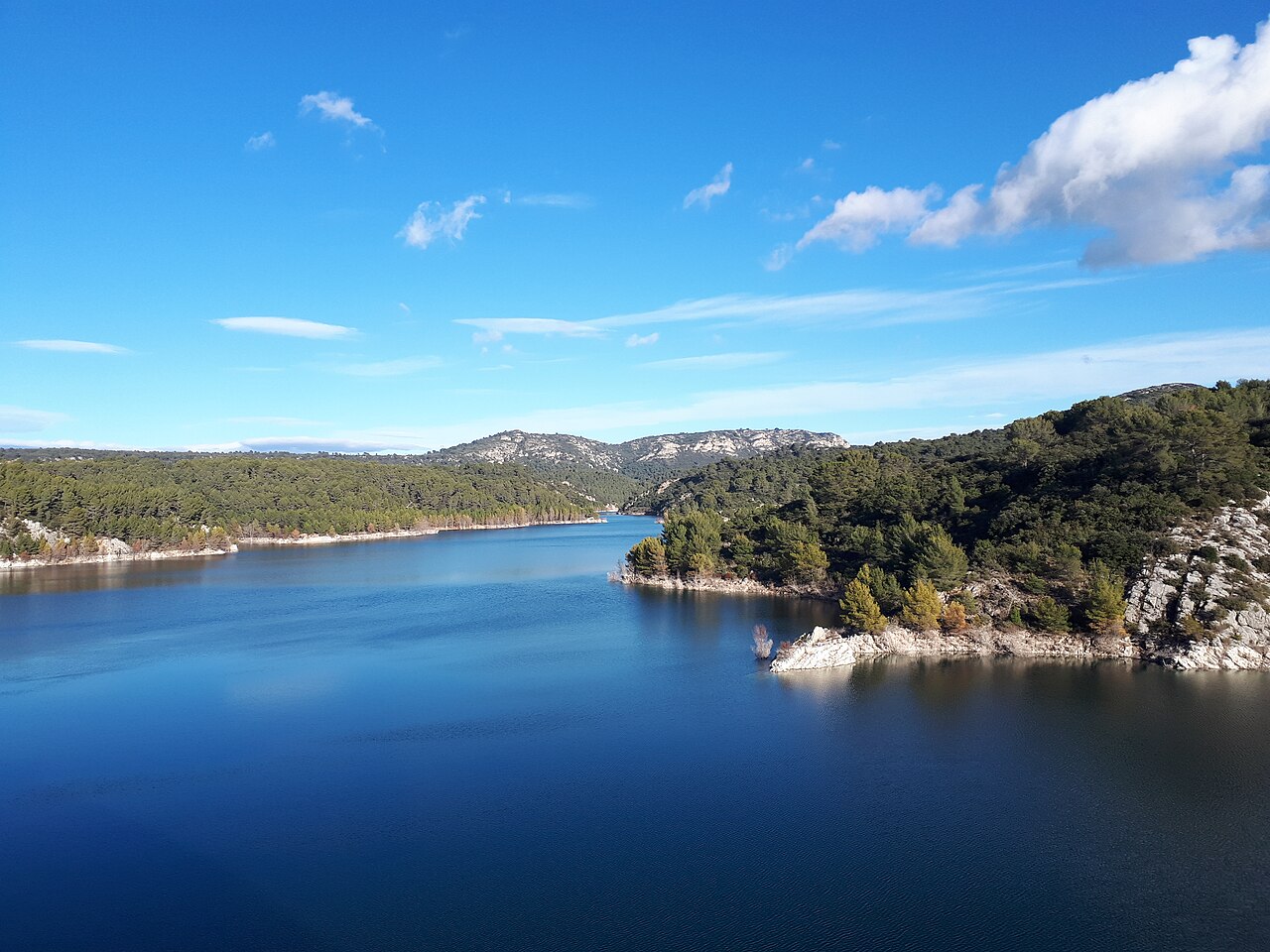 Le lac de Bimont sous un ciel bleu : eau profonde et sombre, berges escarpées couvertes de pins et de calcaire blanc, collines de la Sainte-Victoire à l'horizon