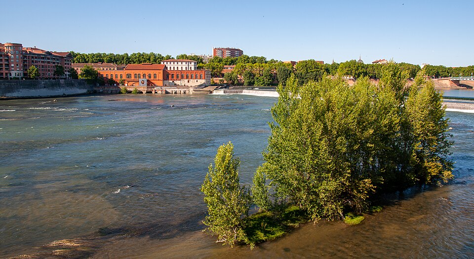 La Garonne à Toulouse au niveau de la chaussée du Bazacle : le fleuve large et peu profond, la chaussée et l'usine hydroélectrique en brique rouge sur la rive gauche, une île de peupliers au milieu du lit