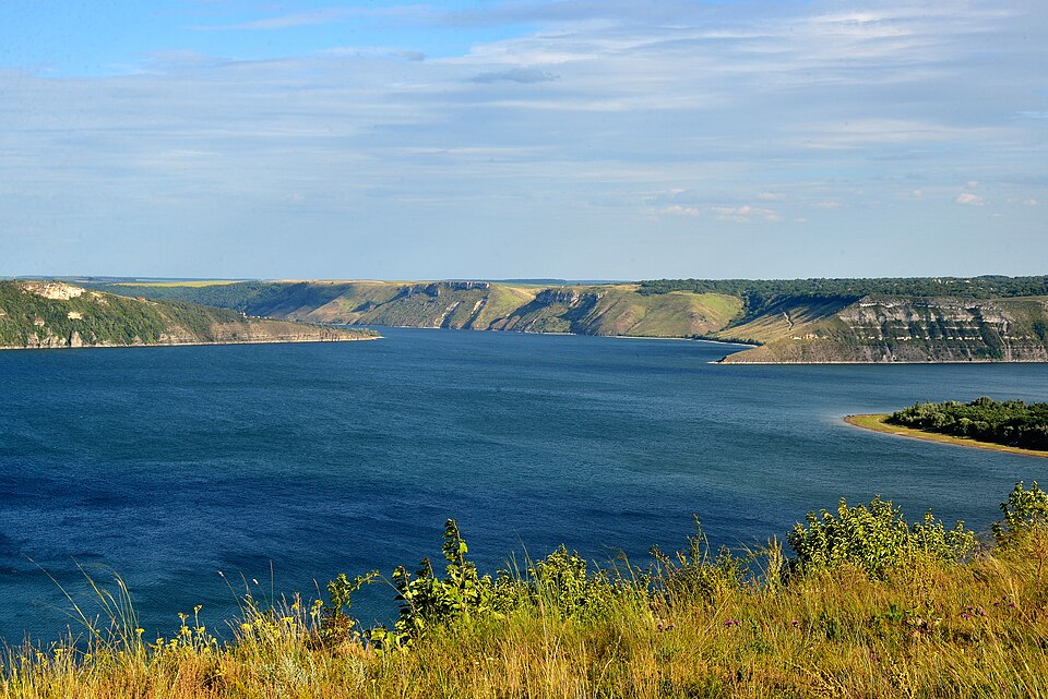 Дністровське водосховище: широка синя вода в каньйоні між високими берегами з білими вапняковими урвищами, зелений лісистий мис праворуч, трав'янистий схил на передньому плані