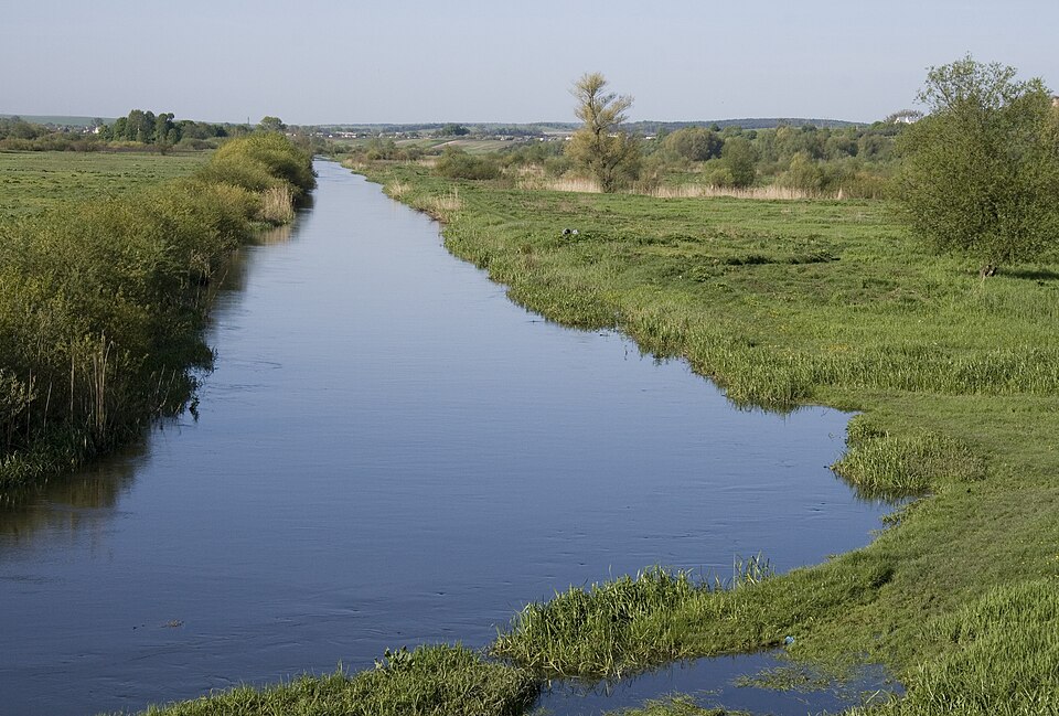 Іква під Дубном: пряме темне русло серед заплавних лук, верболіз і очерет на лівому березі, скошений луг праворуч, низькі пагорби на обрії