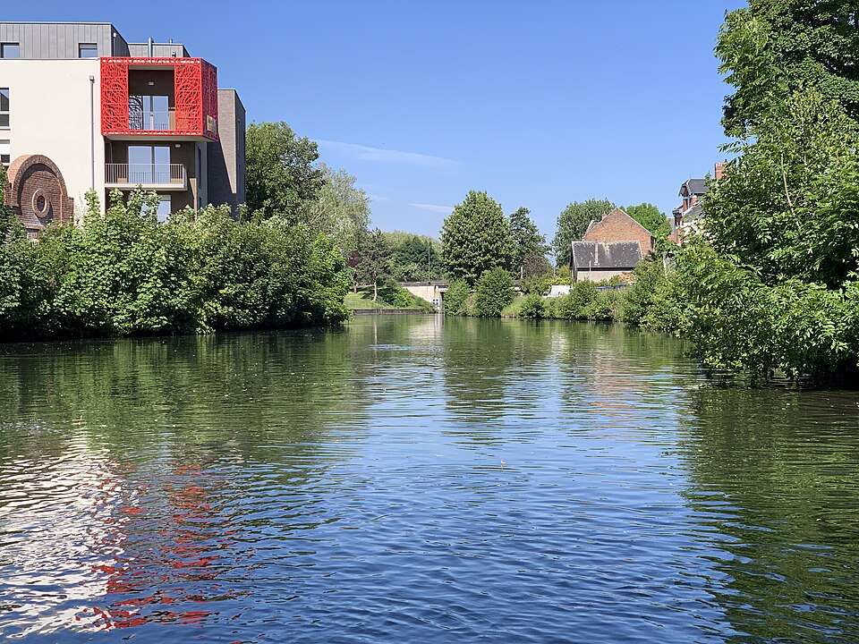 La Somme canalisée à Amiens vue depuis l'eau : le bief large et calme entre deux rives boisées, une écluse au fond, immeubles et maisons de brique sur la rive gauche