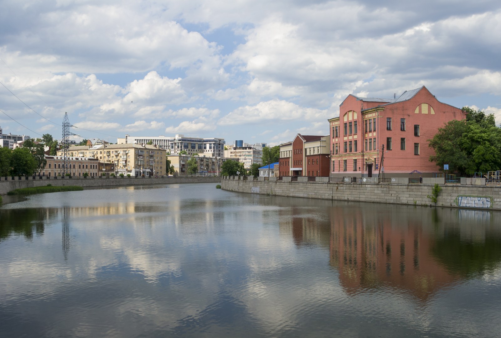 Река Харьков в гранитных набережных: широкая спокойная вода с отражением облаков, кирпичные дома вдоль берега, зелень и городская застройка на горизонте