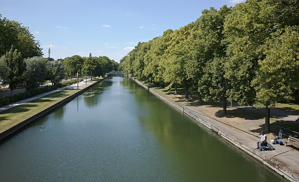 Le canal de la Moyenne-Deûle à Lille vu depuis un pont : le bief rectiligne entre deux berges maçonnées, doubles rangées de platanes, un pêcheur assis avec son matériel sur le quai de droite