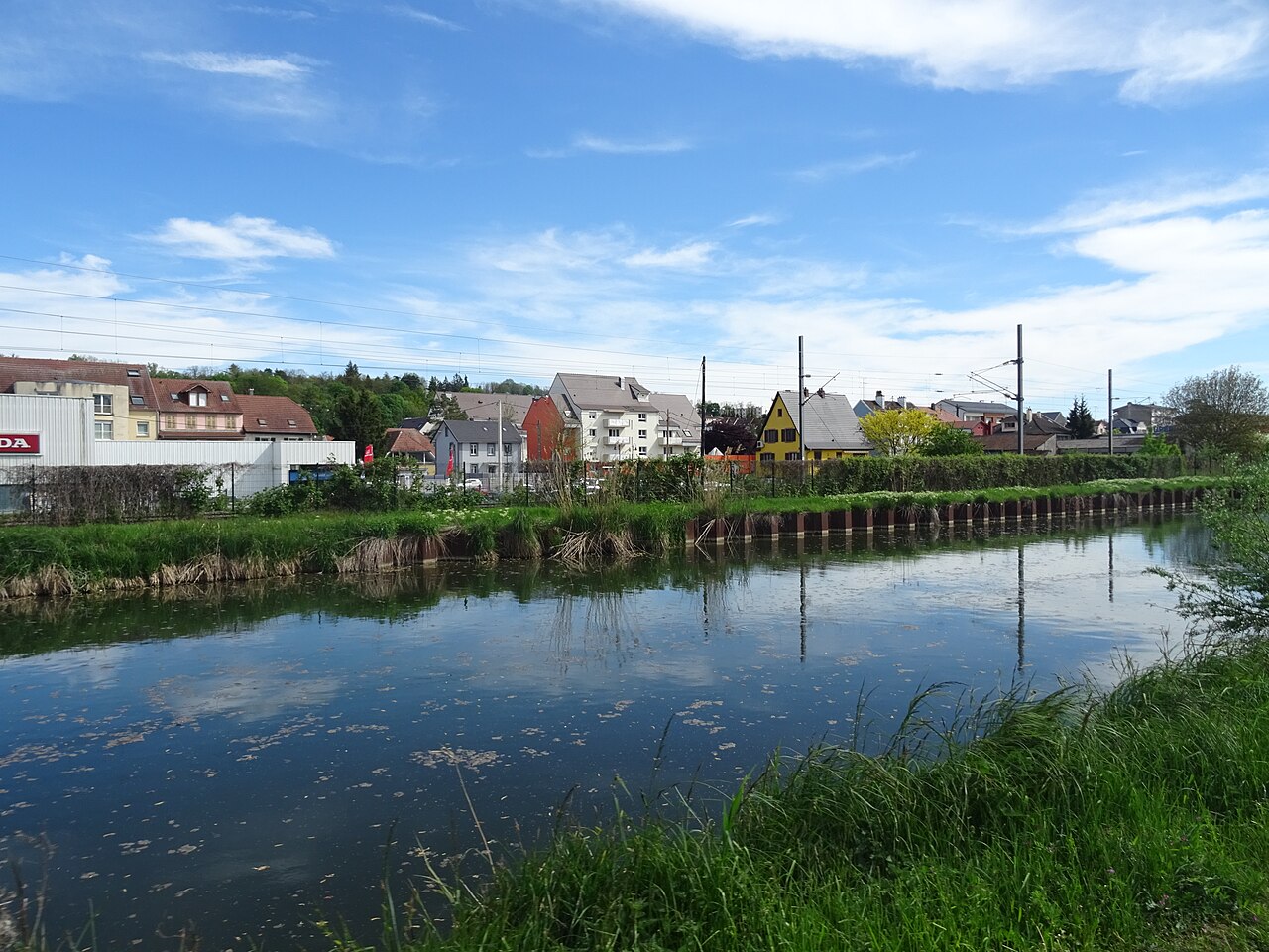 Le canal du Rhône au Rhin à Mulhouse : eau plate et calme reflétant le ciel, berges enherbées avec palplanches, maisons alsaciennes et caténaires de tramway derrière