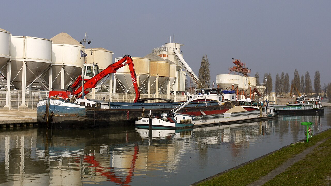 Le canal de l'Ourcq au port de Bondy : péniches de fret amarrées le long du quai, silos et grue derrière, chemin de halage enherbé au premier plan