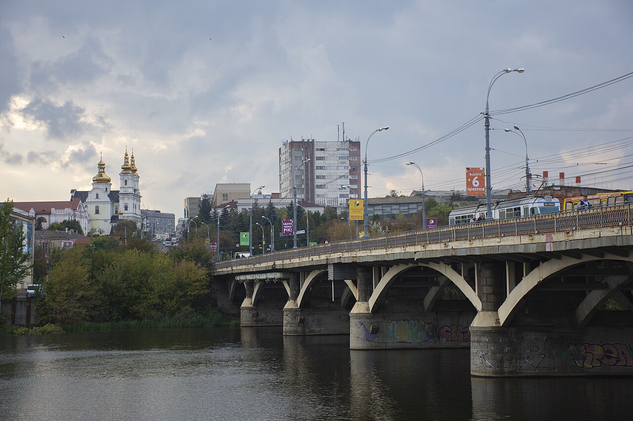 Південний Буг біля Вінниці восени: спокійна вода з відображенням лісистого схилу в жовтій і зеленій листві, очерет уздовж берега, невеликий причал праворуч