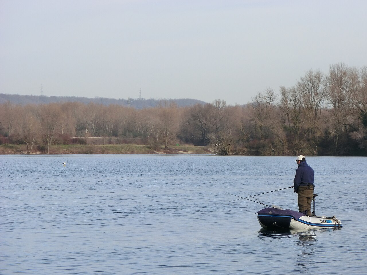 Un pêcheur debout dans une barque équipée d'un moteur électrique sur le lac des Eaux Bleues, eau calme et large, rive boisée d'arbres nus et collines à l'horizon