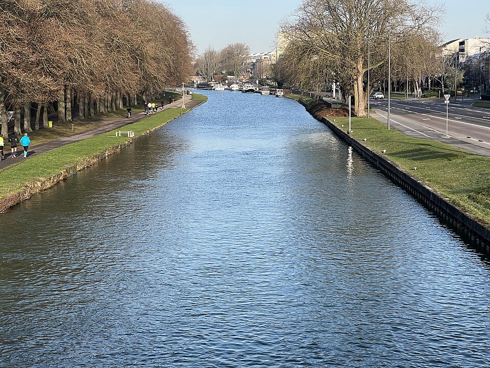 Le canal de l'Aisne à la Marne à Reims vu depuis un pont : le bief large et rectiligne entre deux berges de palplanches, platanes et chemin de halage à gauche, route à droite, bateaux amarrés au loin