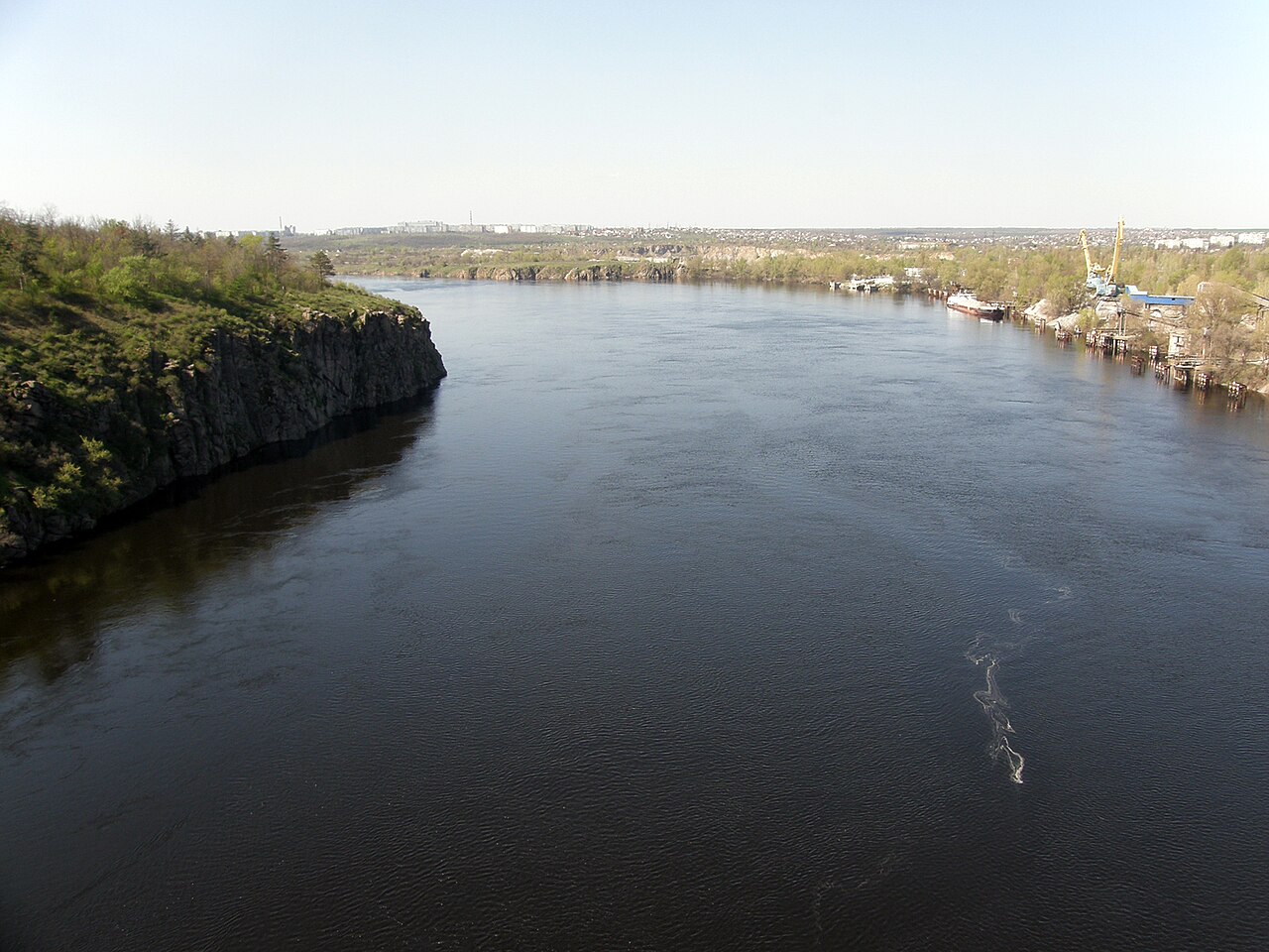 Старий Дніпро вниз від Аркового мосту в Запоріжжі: широка спокійна вода, гранітні скелі лівого берега з зеленню, портовий кран і забудова правого берега вдалині