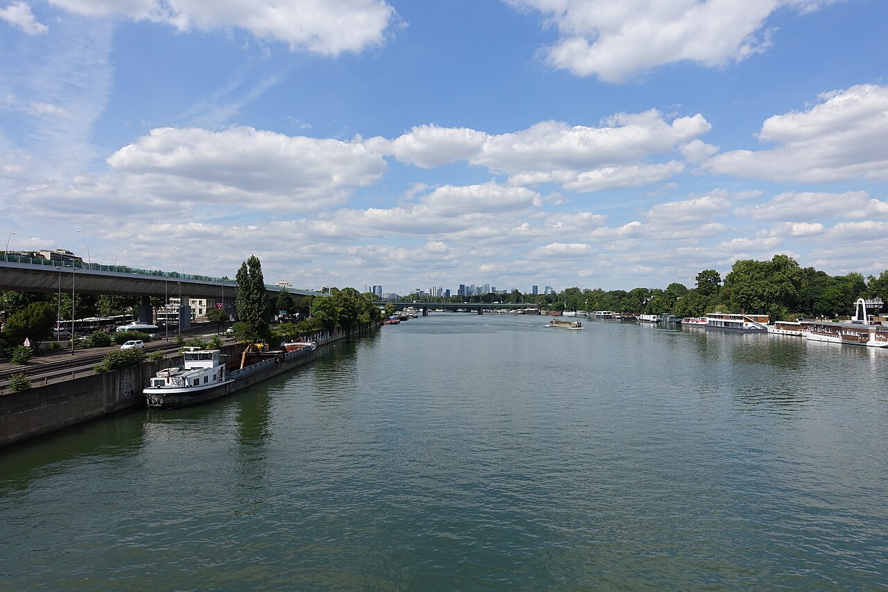 La Seine à Boulogne-Billancourt vue depuis un pont : fleuve large et vert, péniches amarrées le long du quai de gauche, bateaux-logements à droite, tours de La Défense à l'horizon