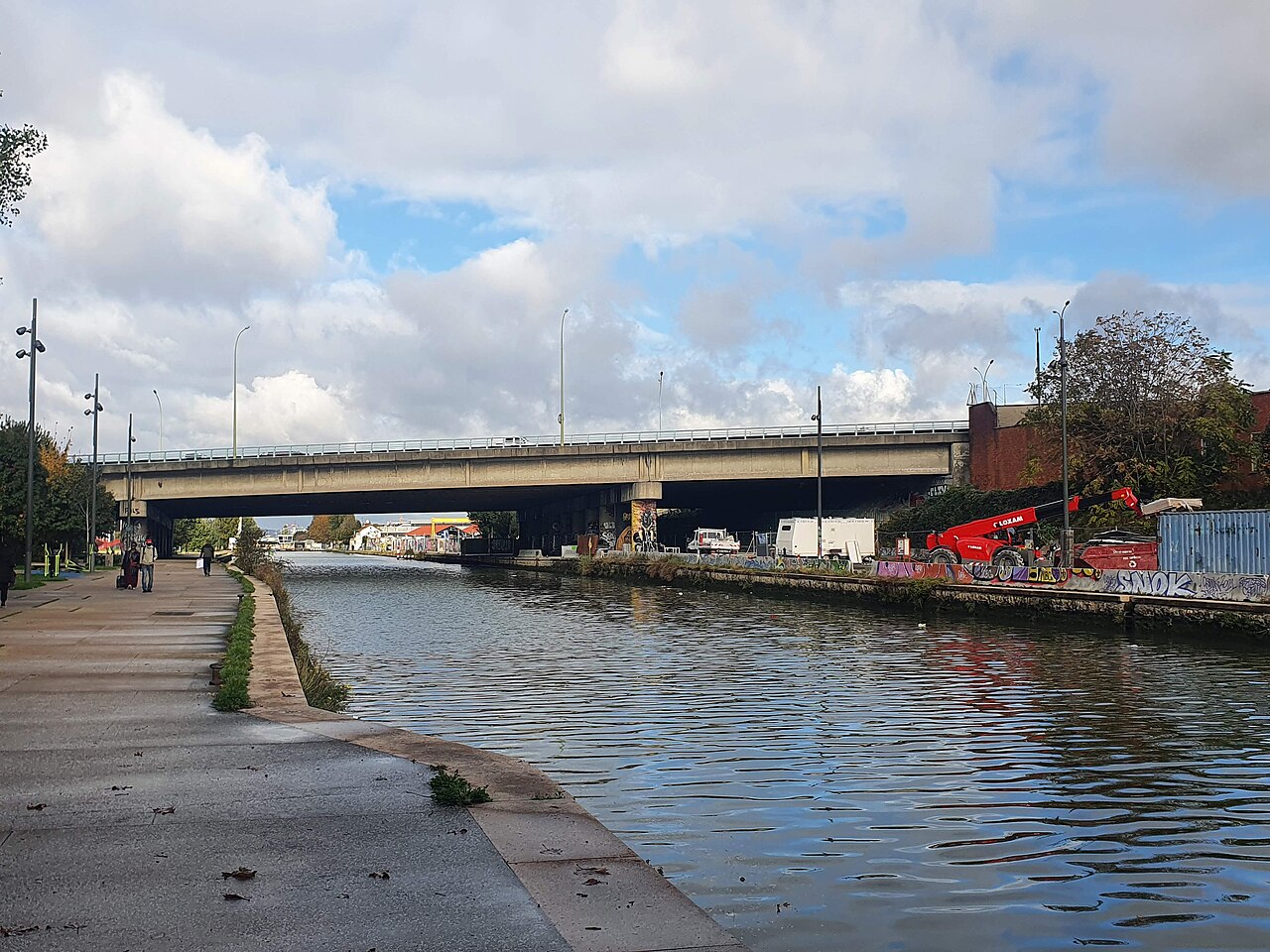 Le canal Saint-Denis vu depuis le quai : eau plate bordée d'un large chemin de halage pavé, un pont routier en béton, rive opposée industrielle avec grue et palissades taguées