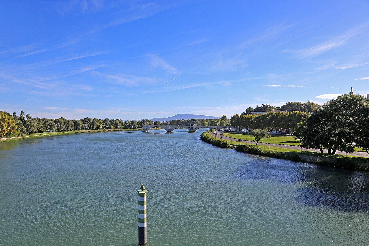 Le Rhône à Avignon vu depuis un pont : le fleuve large et calme, le pont Saint-Bénézet et ses arches au fond, berges arborées des deux côtés, silhouette du Mont Ventoux à l'horizon