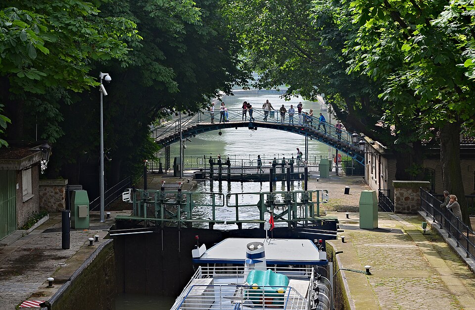 Le canal Saint-Martin à Paris aux écluses des Récollets : le bief entre deux quais pavés, une passerelle en fer arquée au-dessus de l'eau, platanes des deux côtés, une péniche dans le sas