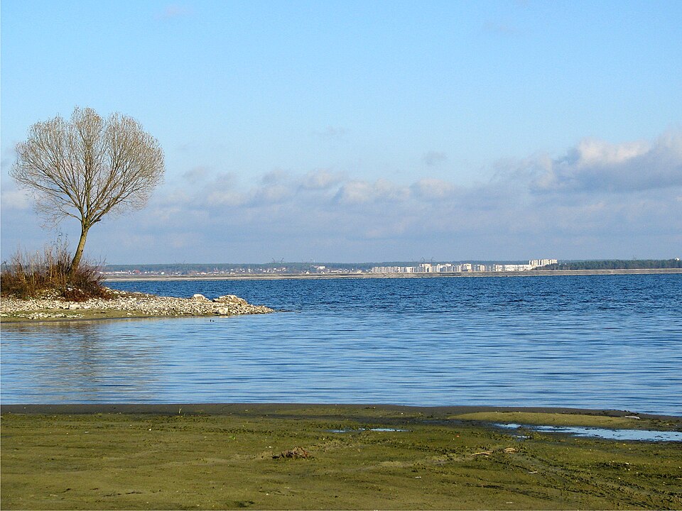 Водосховище-охолоджувач біля Нетішина: широка синя вода під високим небом, гола верба на кам'янистій косі ліворуч, багатоповерхівки міста смугою на тому березі