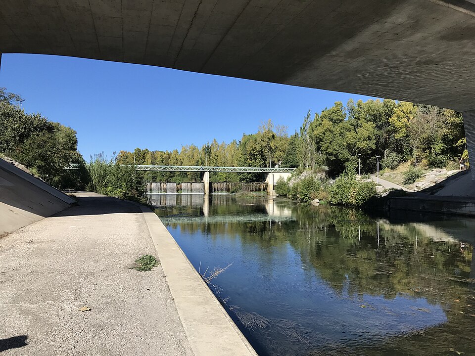 Le Lez à Montpellier vu depuis le dessous d'un pont routier en béton : la rivière calme entre des berges bétonnées, un seuil barré d'une passerelle métallique verte au fond, végétation méditerranéenne sur les deux rives