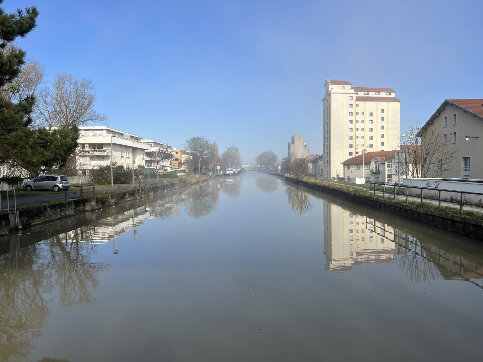 Le canal de la Marne au Rhin à Nancy en hiver : une eau plate et immobile entre deux berges droites, immeubles et maisons se reflétant dans la surface
